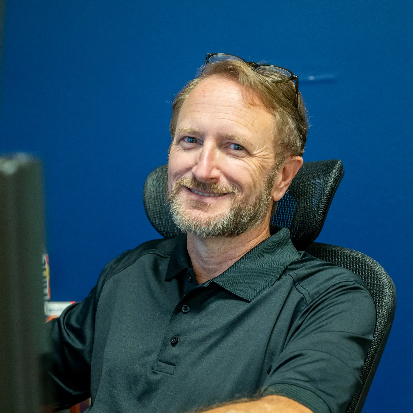 Smiling middle-aged man with beard sitting at desk in office wearing a black polo shirt and glasses on head.