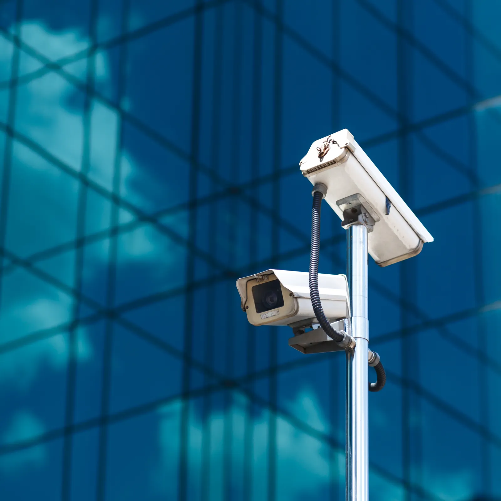 Two white security cameras mounted on a pole in front of a reflective glass building with blue sky reflected.
